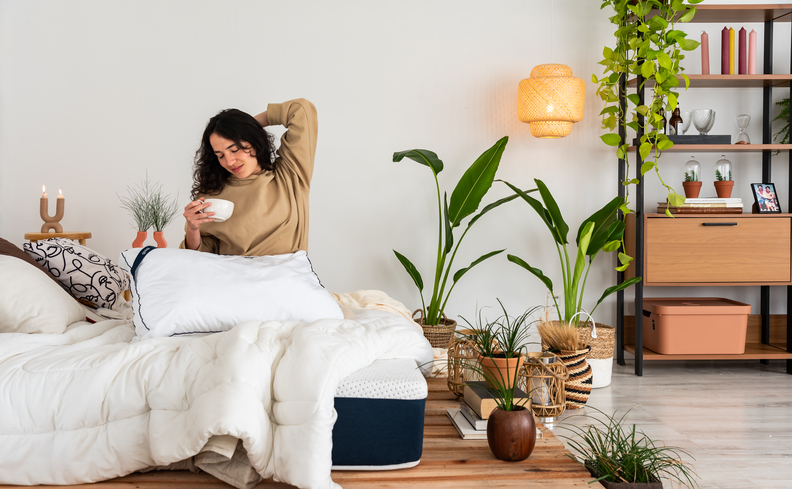 dor nas costas o que pode ser com uma mulher sentada na cama se espreguiçando segurando uma xícara de café com edredom branco, travesseiros, além de estante decorativa e vasos com plantas.