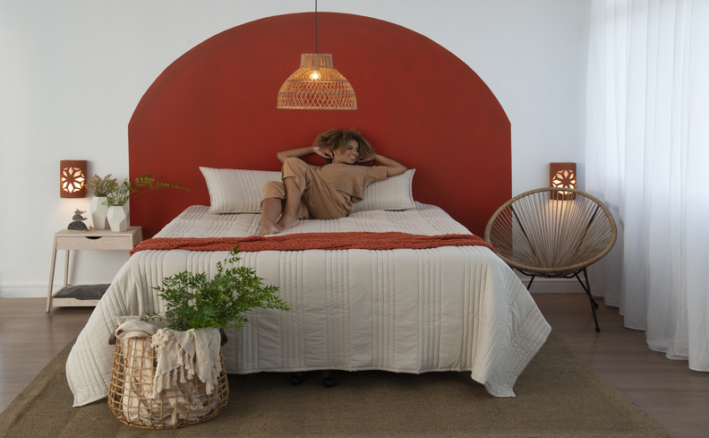 mulher deitada na cama com dor nas costas em um quarto com cama, colchão, edredom branco e peseira laranja, além de dois travesseiros, mesa de cabeceira, poltrona e lustre pendente.