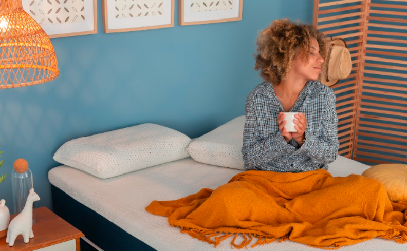 curiosidades sobre cronotipo com mulher sentada na cama segurando uma xícara de café, com uma manta laranja no quarto com quadro decorativo, travesseiros e lustre pendente.