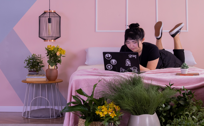 tipos de colchão de casal, mulher deitada na cama usando o notebook, itens de decoração de vaso com plantas, pendentes e outros objetos.