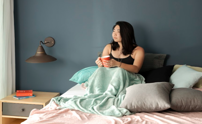 dicas para dormir melhor com uma pessoa sentada na cama segurando uma xícara de chá, com cobertores e travesseiros