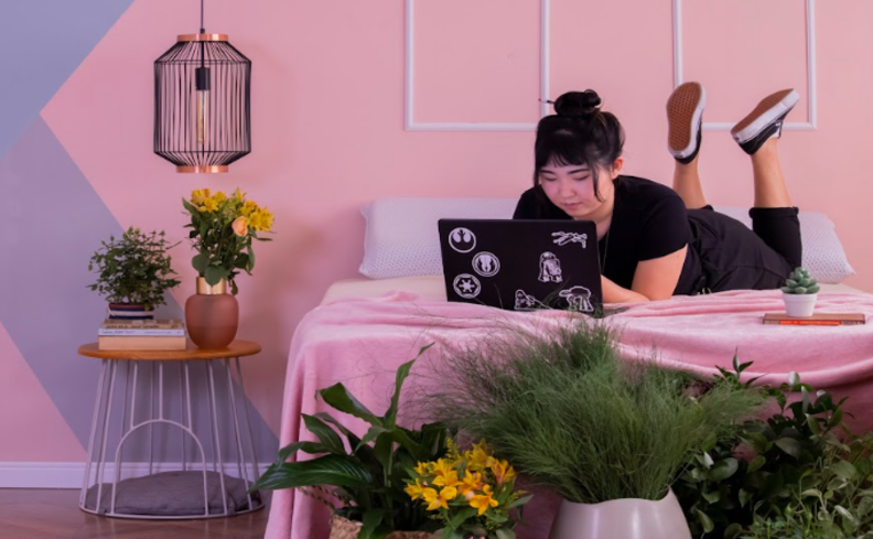 dormir pouco faz mal para a saúde, pessoa deitada na cama mexendo no notebook em um quarto com cama, pendente, mesa de cabeceira e itens decorativos