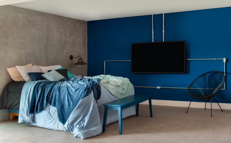 dormir tarde faz mal com quarto com cama, roupa de cama azul com travesseiros, banco, poltrona e televisão