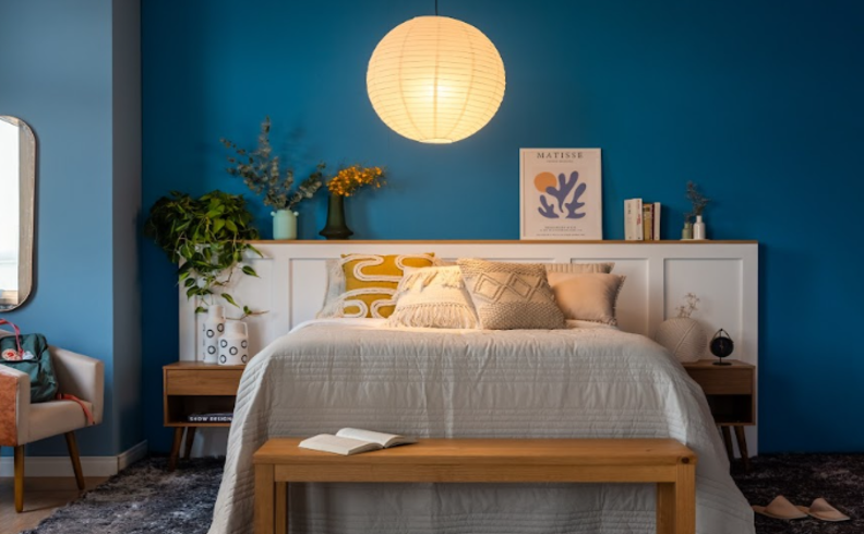 quarto com iluminação noturna, paredes azuis, cama posta com edredom e almofadas, quadros e vasos com plantas decorativos.