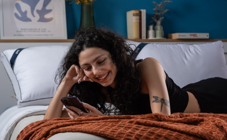mulher deitada de bruços na cama mexendo no celular procurando saber qual o hormônio do sono para conseguir dormir melhor.
