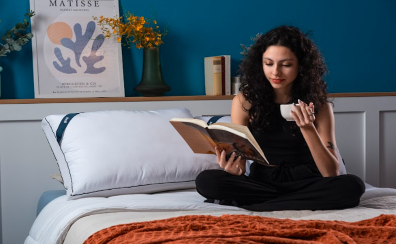 mulher sentada na cama lendo sobre as bebidas com cafeína e segurando uma xícara.