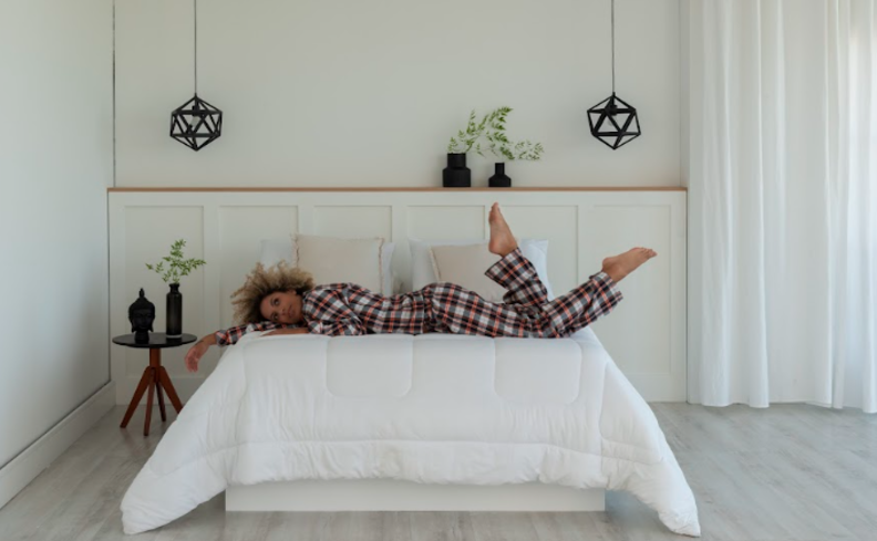 mulher deitada na cama em um quarto com a decoração toda branca, com luzes pendentes e uma mesa de cabeceira com vasos de plantas pensando em como estimular o hormônio do sono.