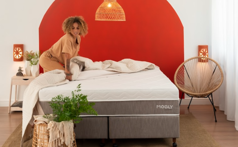 como eliminar ácaros no colchão e travesseiro com mulher trocando a roupa de cama em um quarto com edredom, além de mesa de cabeceira, poltrona e itens decorativos.