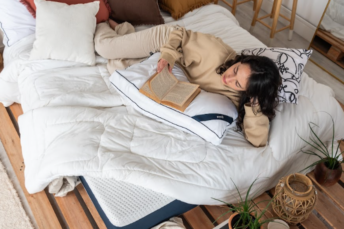 dicas para começar a ler antes de dormir com uma mulher deitada apoiada no braço lendo um livro em uma cama confortável com um travesseiro e edredom.