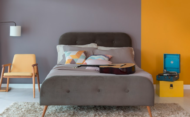 como dormir rápido em um quarto com cama com pés palito, cabeceira cinza, poltrona, vitrola, tapete e diversos travesseiros e almofadas.