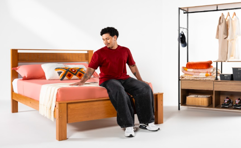 saber se dormir depois do almoço faz mal ou não é importante para melhorar a qualidade do sono, homem sentado na cama em um quarto com cama e arara de roupas.