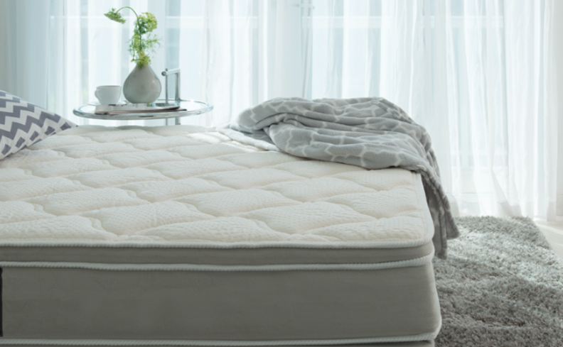 dicas para saber o que é mola ensacada em um quarto com colchão branco, tapete cinza, cortinas, mesa de cabeceira com vaso e xícara, além de dois travesseiros e edredom na cama.