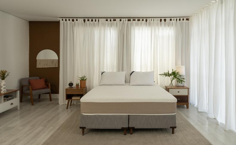 colchão bom no quarto com base box de cama, tapete, mesas de cabeceira, poltrona, vasos com flores decorativos, almofadas e dois travesseiros.