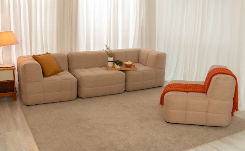 sofá confortável para sala, com 4 módulos e um usado como pufe, além de tapete, luminária e mesa lateral.