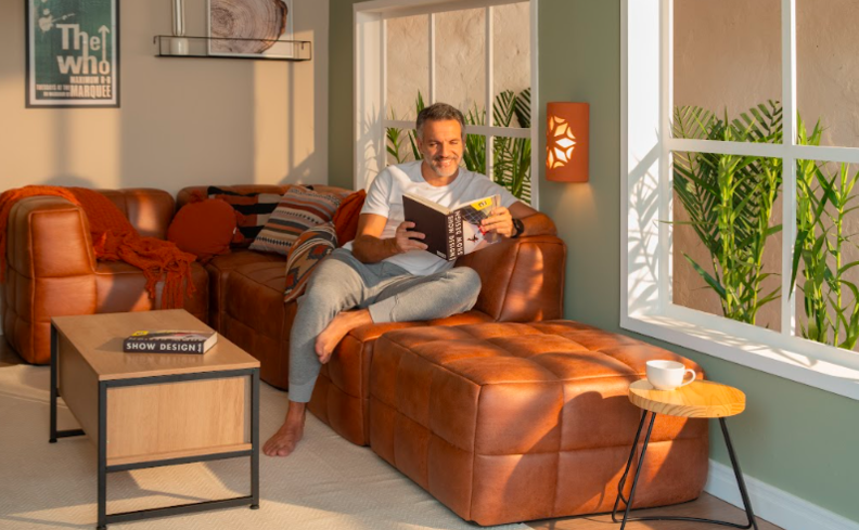 sofá em módulos na cor marrom na sala de estar, além de mesas de centro em madeira e homem sentado no sofá lendo um livro.
