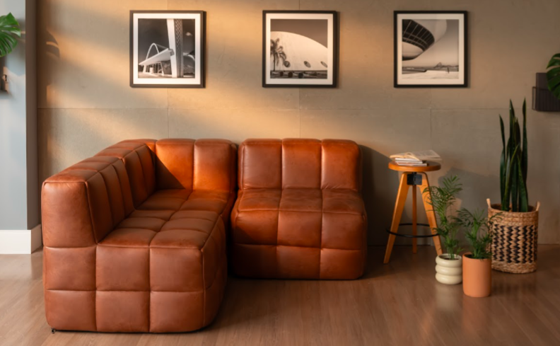 sala de tv pequena com sofá modular marrom em formato L, banquinho de madeira e três vasos com plantas.