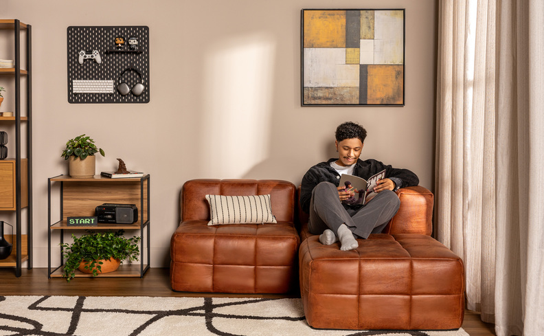 sala de tv pequena com sofá em modulos com um homem sentado lendo um livro em um sofá marrom, tapete, quadro decorativo e mesa de apoio quadrada ao lado.