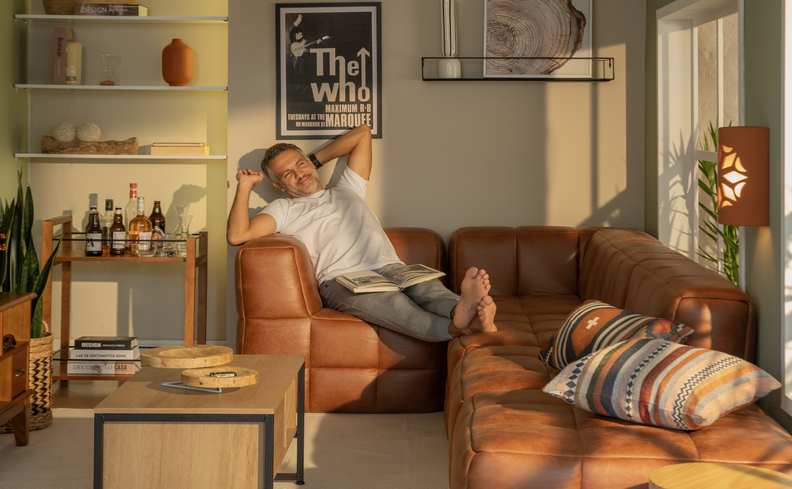 sofá modular marrom na sala de tv pequena com homem se espreguiçando no sofá, além de diversas almofadas, mesa de centro e várias estantes nas paredes.