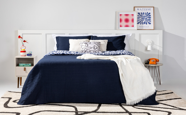 colchão macio e confortável em um quarto com roupa de cama em tons de azul e branco, com diversas almofadas e travesseiros para saber como fazer a cama igual a de hotel, além de mesas de cabeceira e tapete.