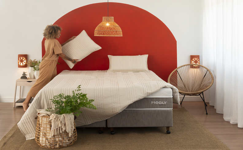 como fazer a cama igual a de hotel com mulher arrumando os travesseiros na cama com base box bipartida, luminária, parede com detalhe laranja e roupa de cama branca.