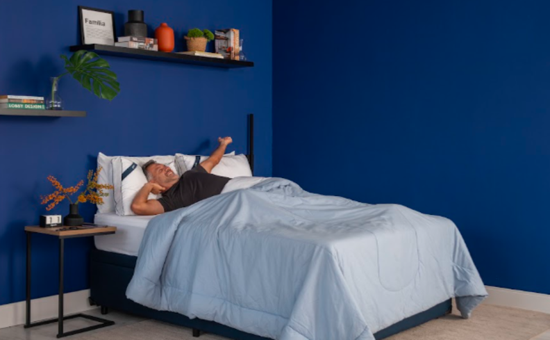 homem deitado em uma cama com um bom colchão e travesseiro para ter um sono reparador em um quarto com cama, mesa de cabeceira e paredes em tons de azul.