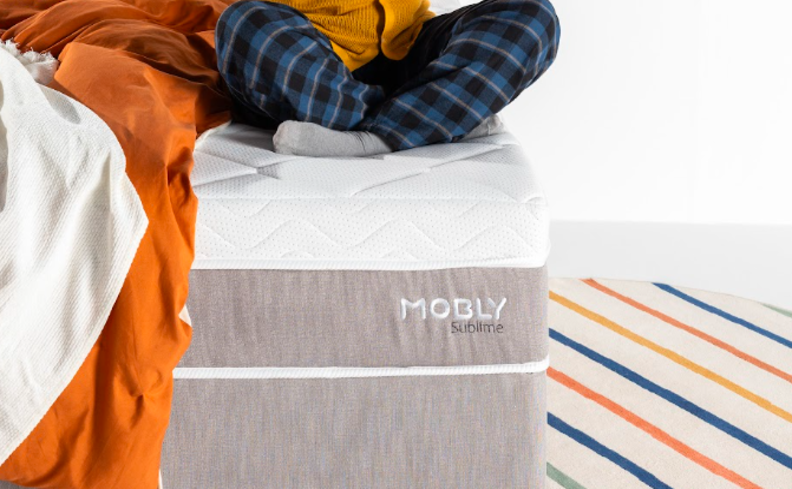 dicas para saber como ter um sono de qualidade com uma mulher sentada em um colchão, com roupas de cama nas cores branca e laranja.